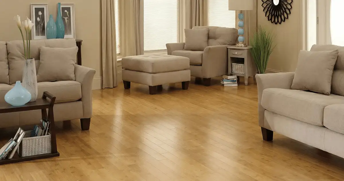 A living room with professionally installed bamboo flooring, beige sofas, a matching ottoman, a side table with a blue lamp, vases, framed pictures, and large windows with blinds and curtains in Glendale, AZ