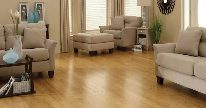 A living room with professionally installed bamboo flooring, beige sofas, a matching ottoman, a side table with a blue lamp, vases, framed pictures, and large windows with blinds and curtains in Glendale, AZ